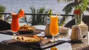 A table set with drinks, plates of food, and a vase with flowers, overlooking the sea. - Lima Satu Resort (Pemenang)