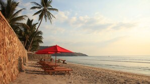 A beach with wooden lounge chairs and a red umbrella, palm trees, and a stone wall. - Lima Satu Resort (Pemenang)