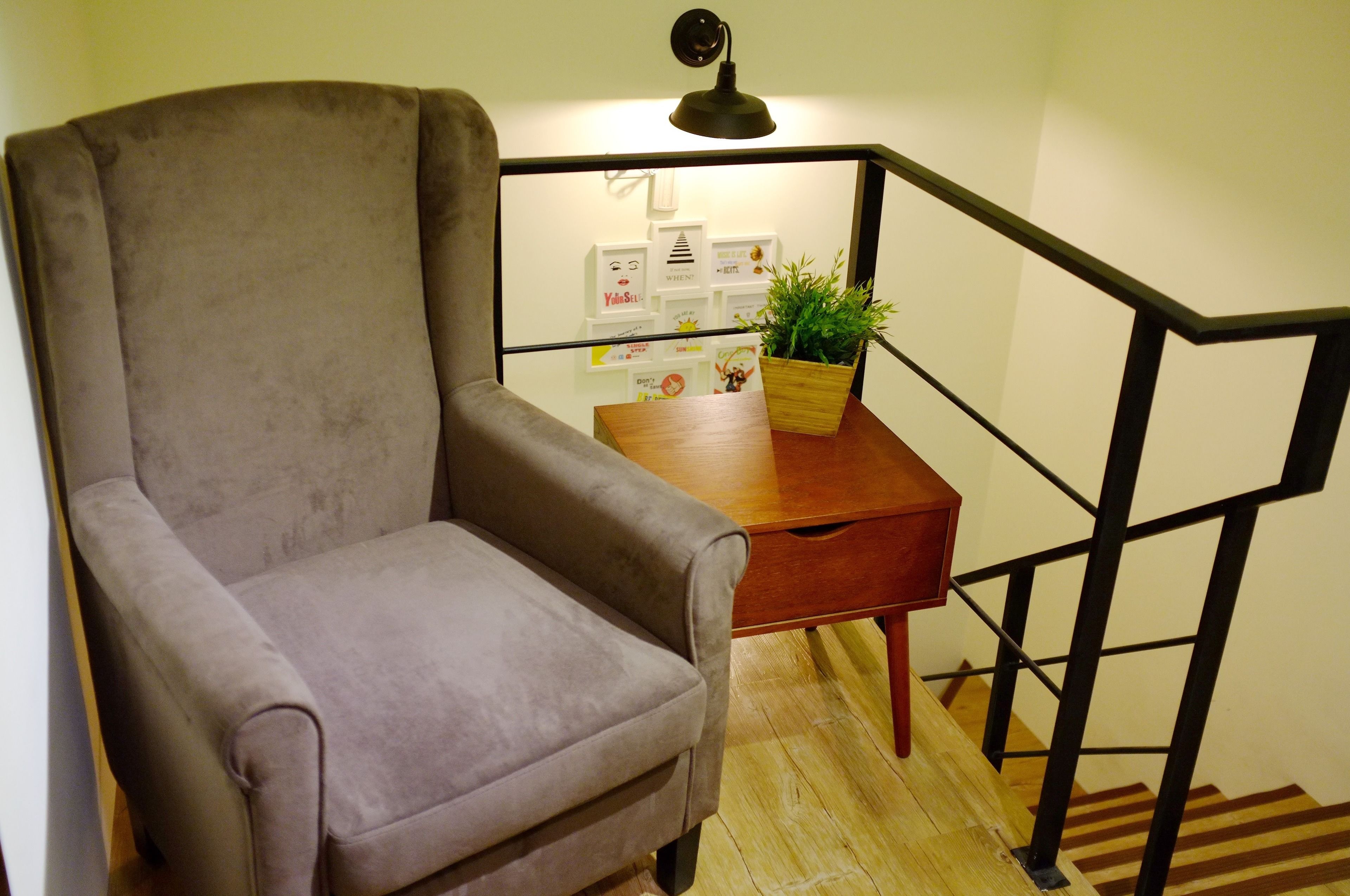 A room with a grey armchair, a wooden side table with a plant, and a staircase with black railings.