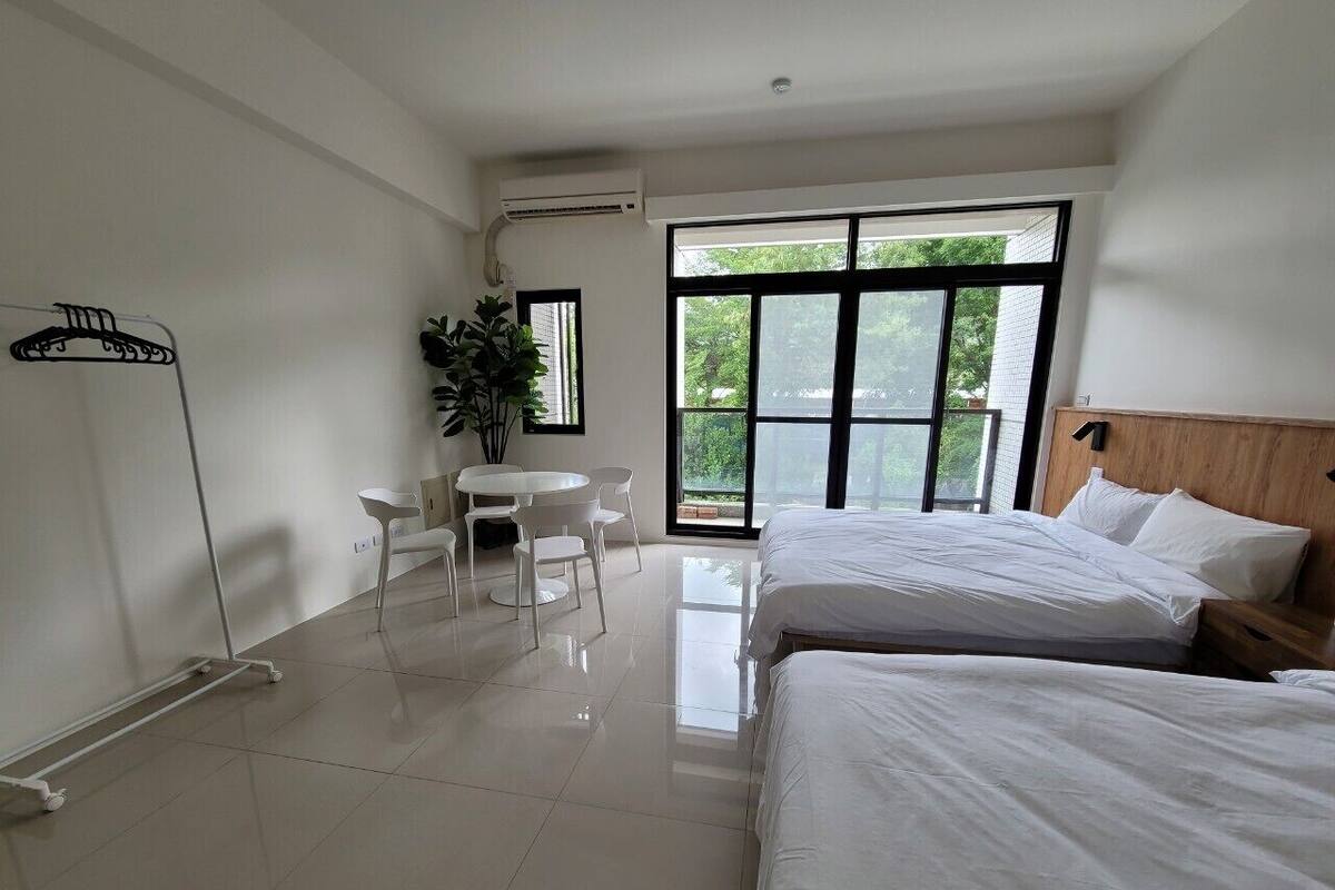 A hotel room with two beds, a small table with chairs, a potted plant, and a large window with a view of greenery.