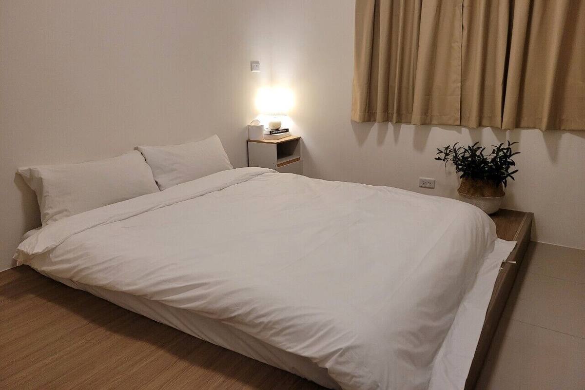 A neatly made bed with white linens in a room with a wooden floor. There is a bedside table with a lamp and a potted plant.