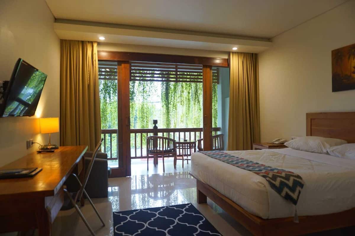 A hotel room with a bed, a desk with a lamp, a chair, and a balcony with a view of greenery.