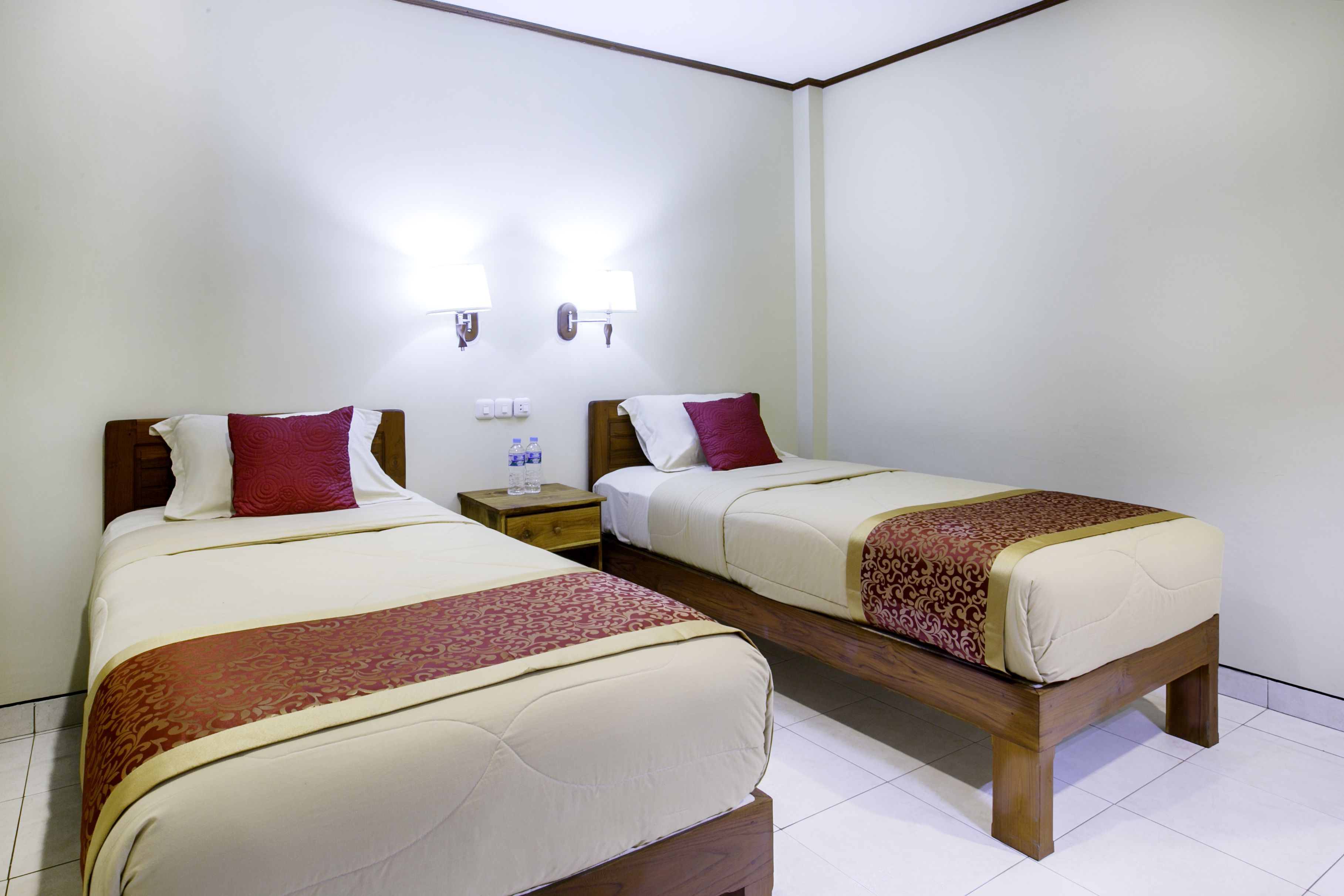 Habitación de hotel con dos camas individuales, cada una con cabecero de madera, ropa de cama blanca y almohadas decorativas rojas. Hay mesitas de noche con lámparas y una pequeña mesa con una botella de agua.