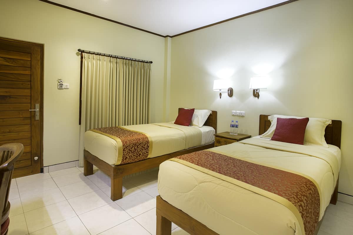 A hotel room with two beds, a wooden door, a curtain, a nightstand, and a wall-mounted lamp.