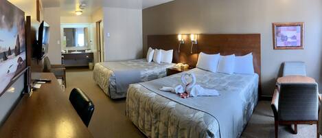 A hotel room with two beds, a desk, a chair, a TV, and a bathroom visible through an open door.