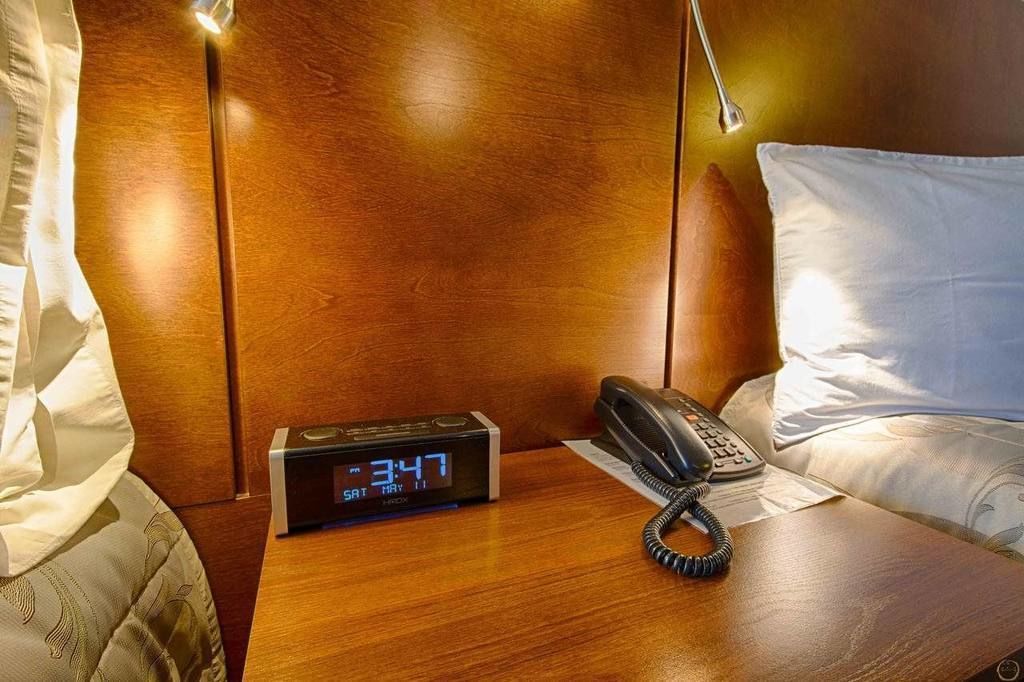 A wooden nightstand with a digital clock displaying 3:47, a telephone, and a pillow.