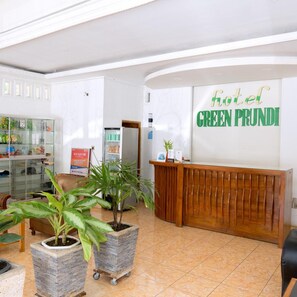 A hotel reception area with a wooden reception desk, a wall-mounted sign reading 'hotel GREEN PRUNDI', and potted plants in the foreground. - Hotel Green Prundi (Labuan Bajo)