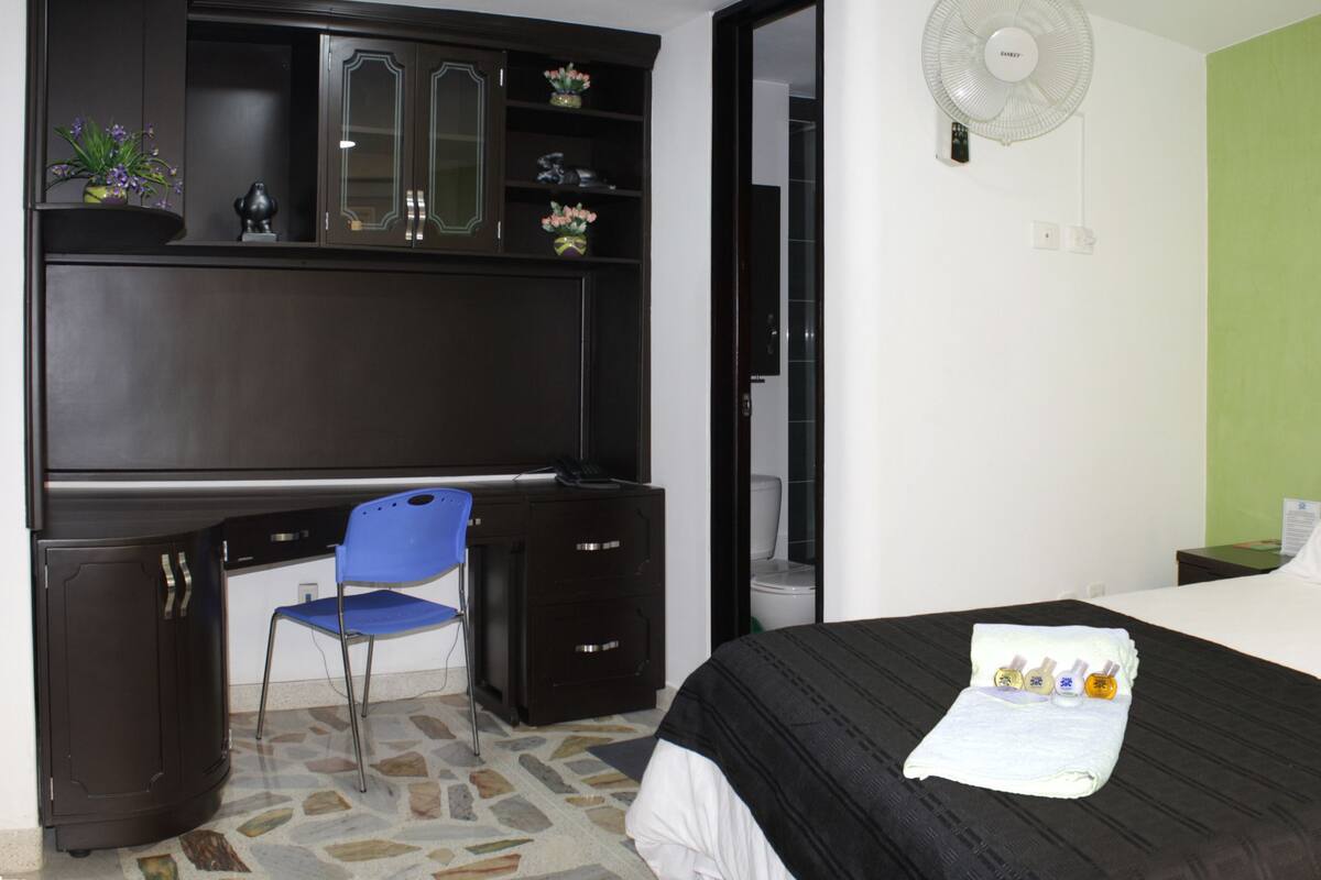 A hotel room with a bed, a desk with a chair, a wardrobe, and a green wall.