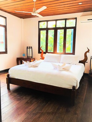 A neatly made bed with white linens, a wooden ceiling fan, and a window with a view of greenery.