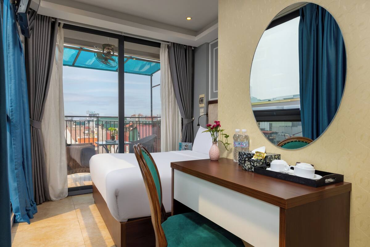 Habitación de hotel con cama, escritorio con bandeja, espejo y vista a la ciudad a través de amplias ventanas.