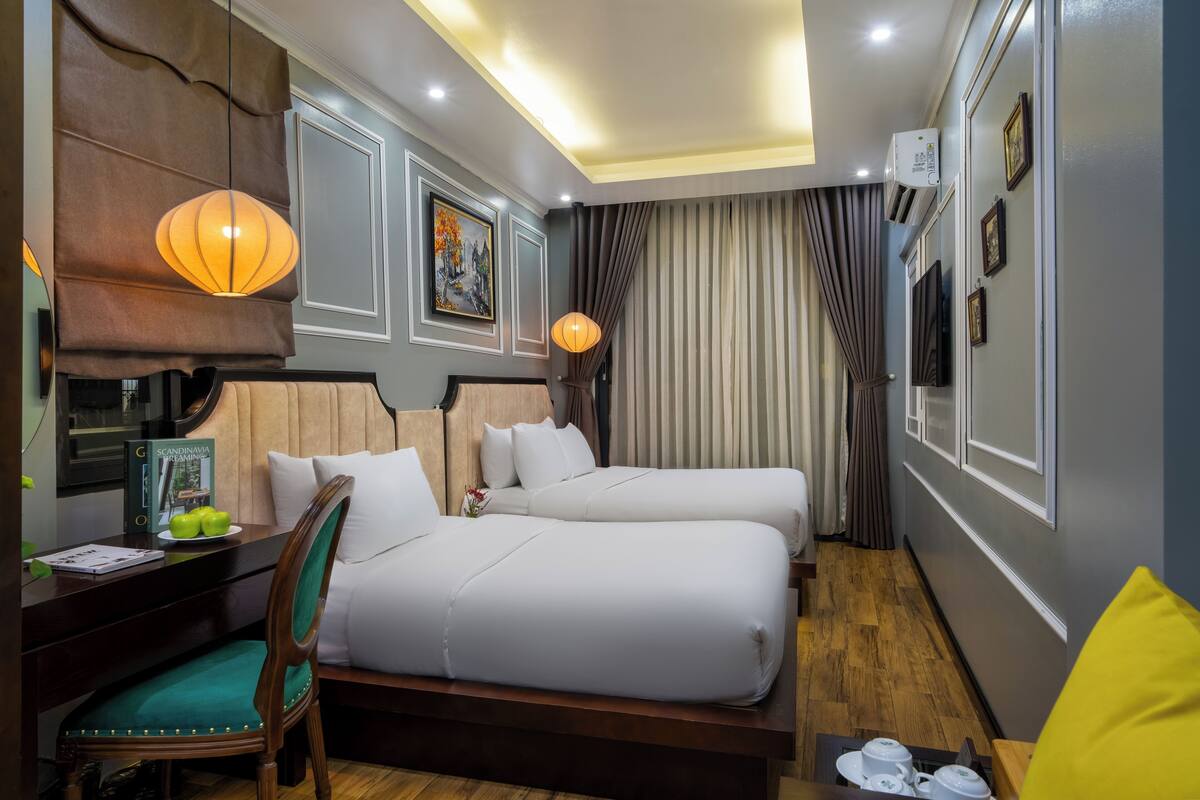 Habitación de hotel con dos camas, un escritorio con silla, una lámpara y un cuadro en la pared.