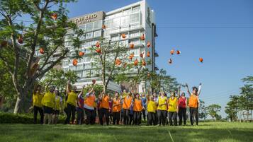 A group of people in yellow and orange vests standing in a grassy area with trees and a building in the background.