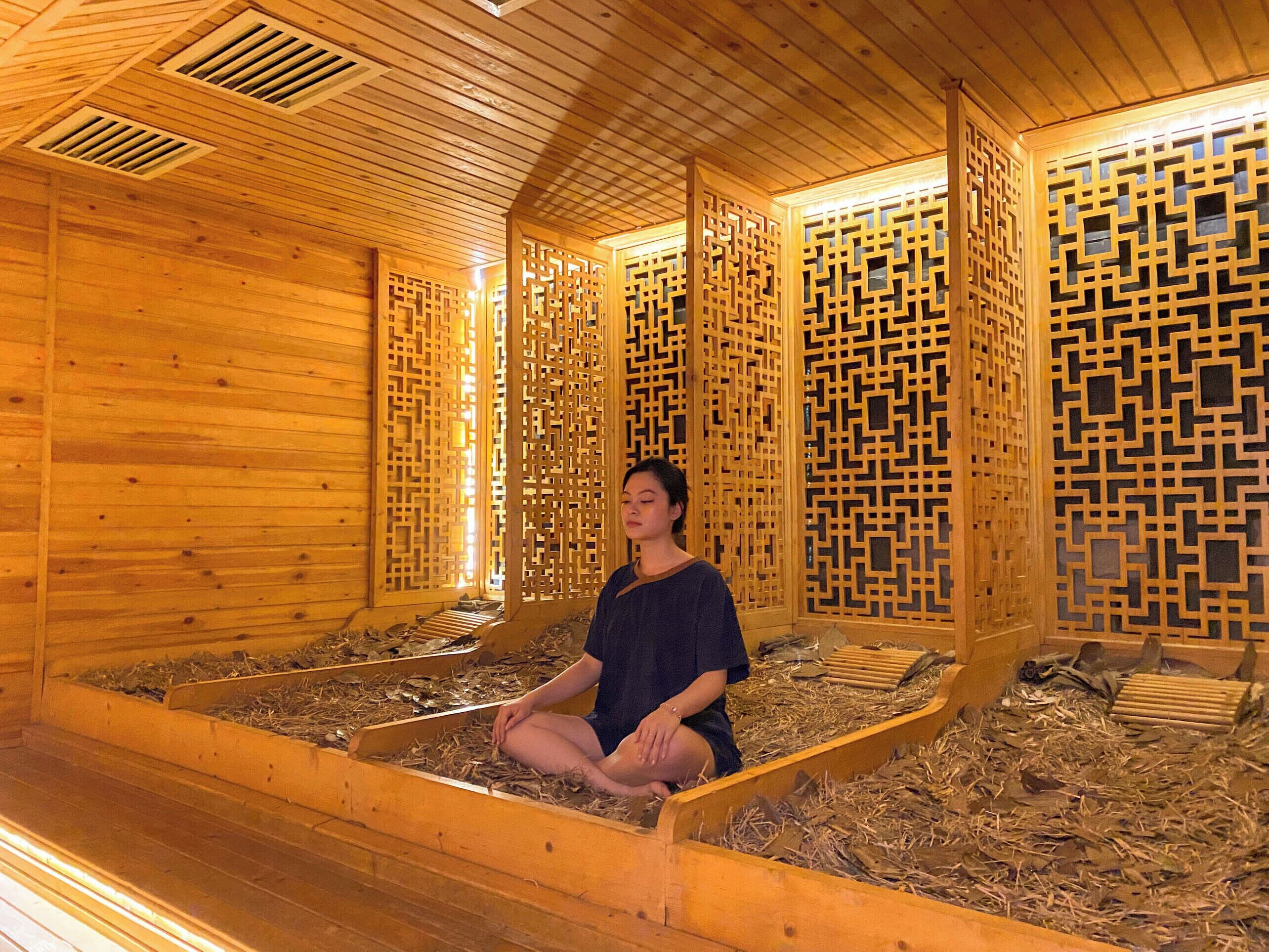 A wooden room with a person sitting on a bench, wooden partitions with geometric patterns, and a sloped ceiling with ventilation.