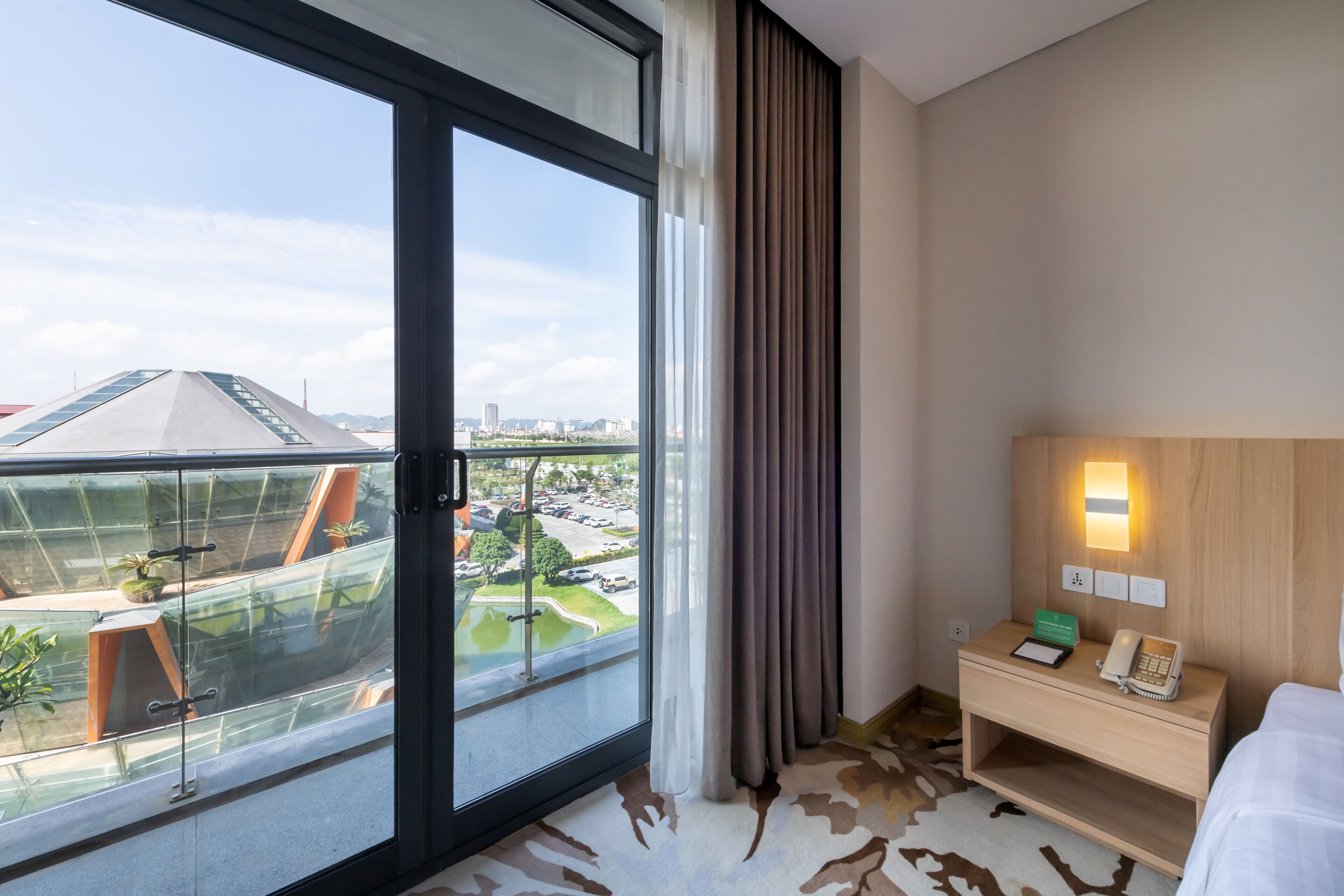 A hotel room with a large sliding glass door, a bed, a bedside table with a lamp, and a view of a cityscape.
