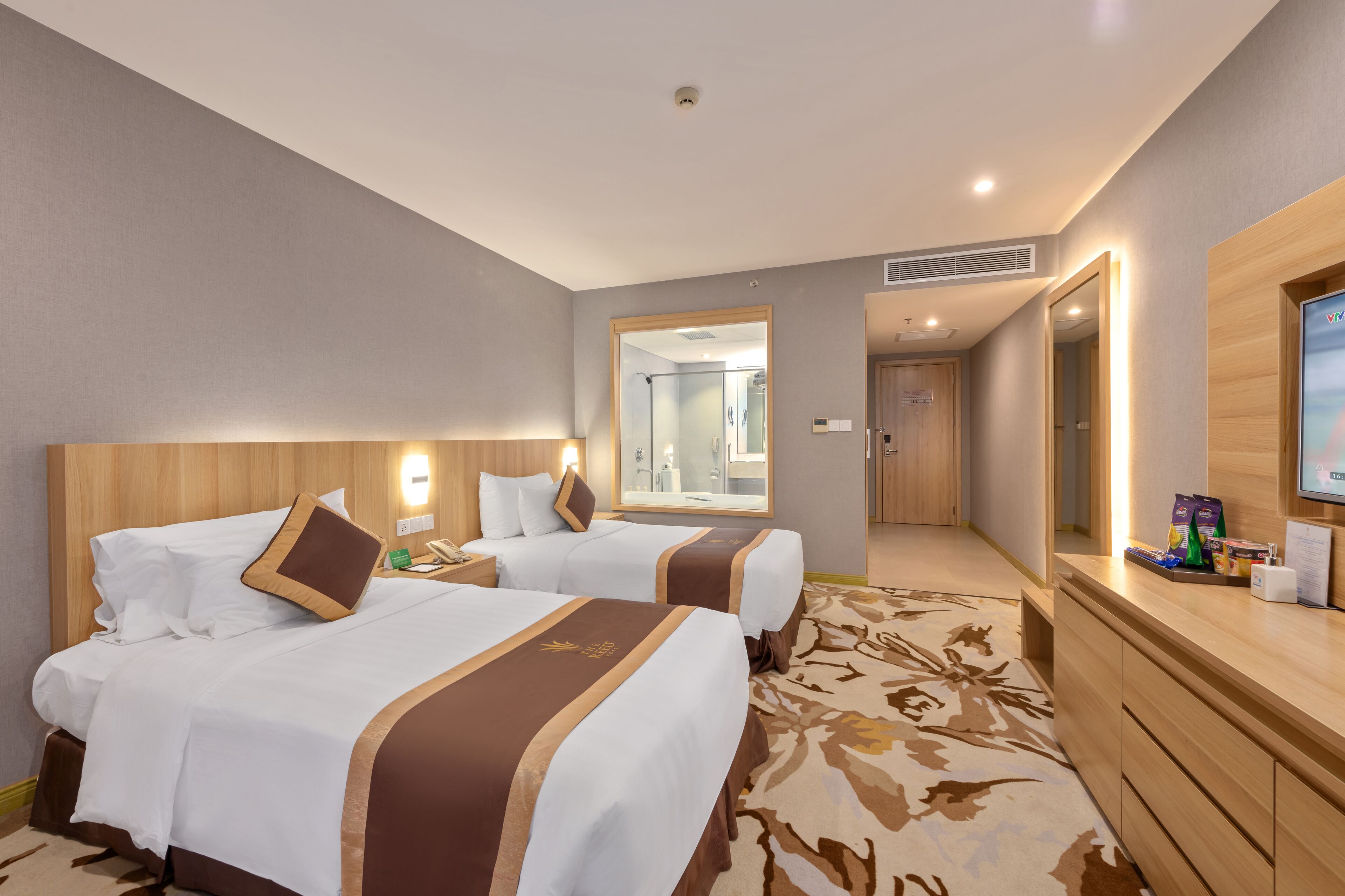 A hotel room with two beds, a wooden headboard, a mirror, a television, and a desk with a computer and some items on it.