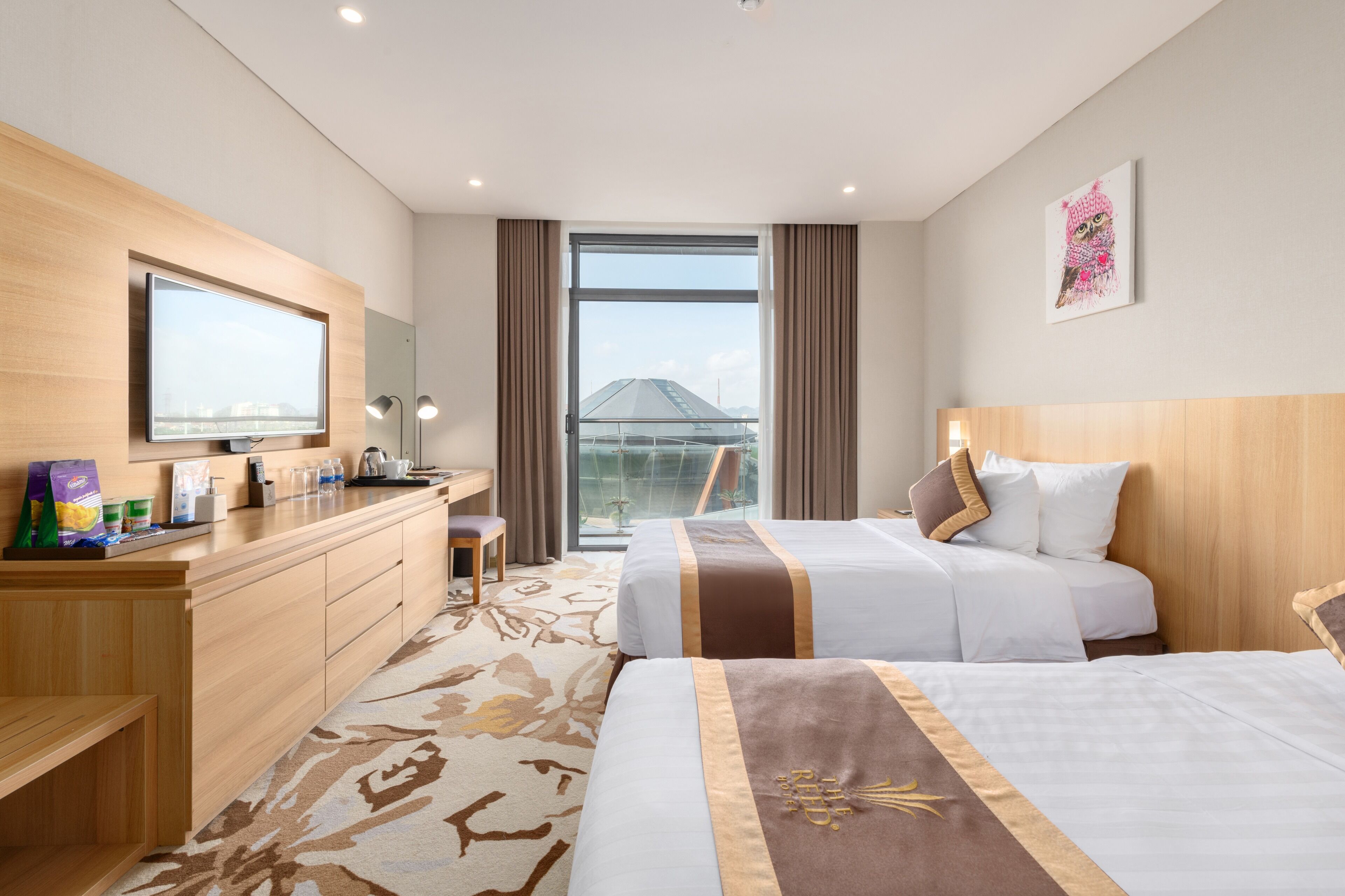 A hotel room with two beds, a desk, a TV, a large window, and a view of a building.