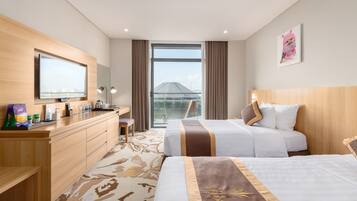 A hotel room with two beds, a desk, a TV, a large window, and a view of a building.