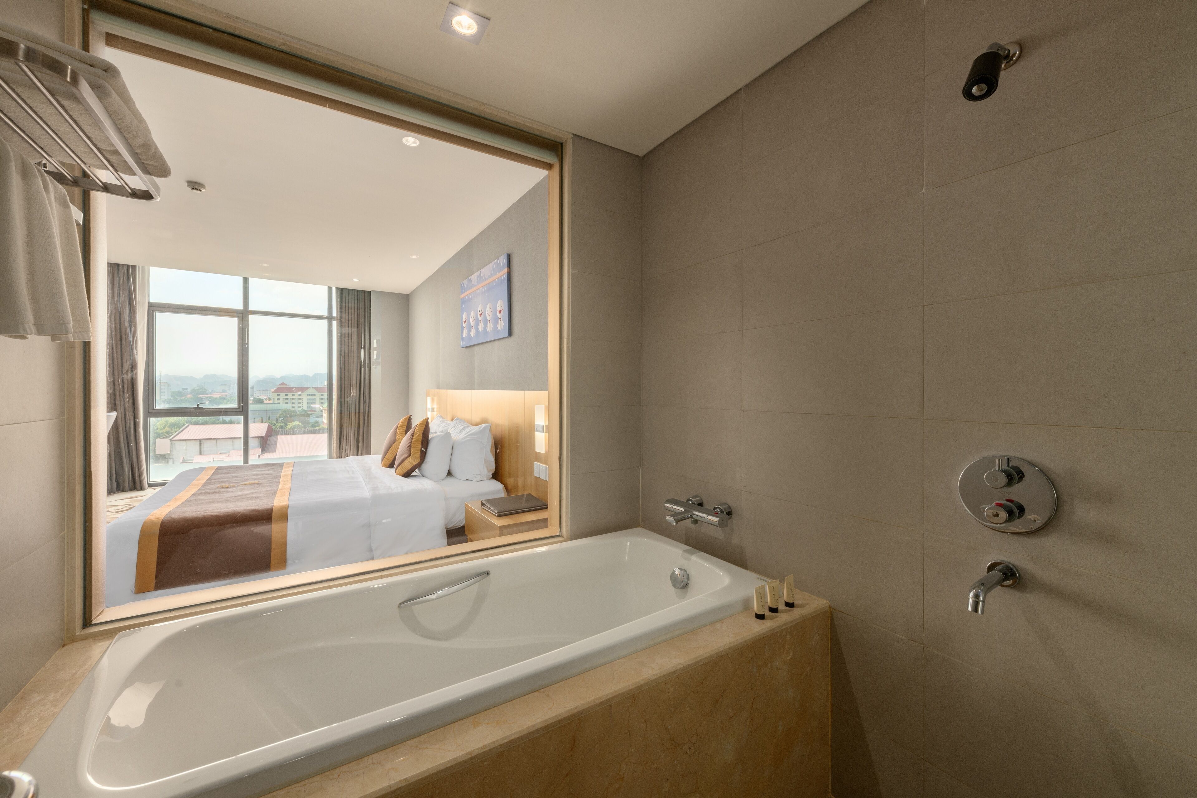 Un moderno baño de hotel con una bañera grande, ducha y espejo que refleja un dormitorio con cama y vista a la ciudad.