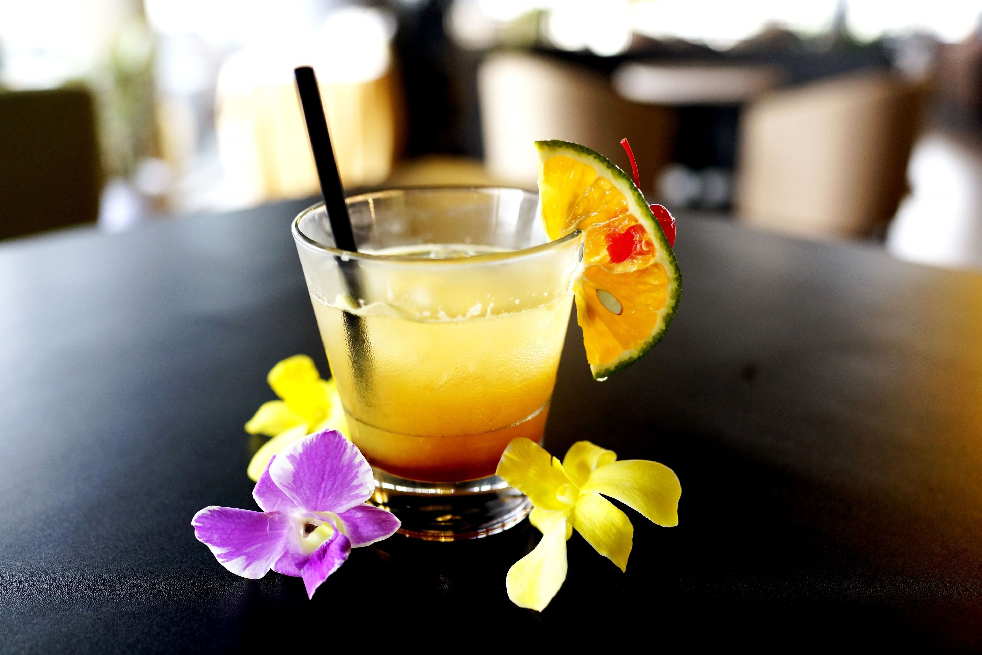 Een glas met een oranje drank, een rietje en een stukje limoen op de rand, met paarse en gele bloemen op een donkere ondergrond.