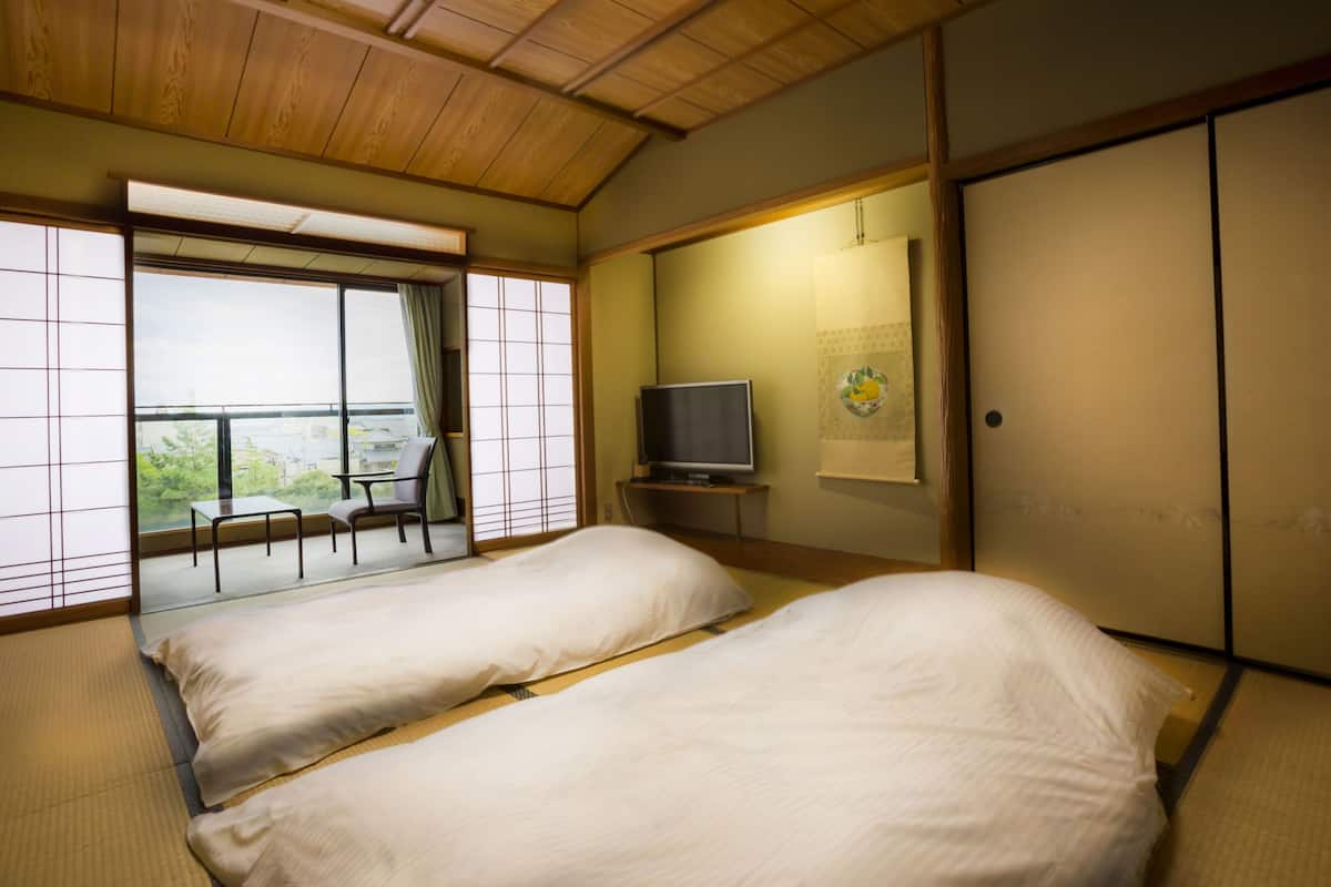 Um quarto japonês tradicional com piso de tatame, uma cama com roupa de cama branca, uma televisão sobre um suporte e uma porta deslizante.
