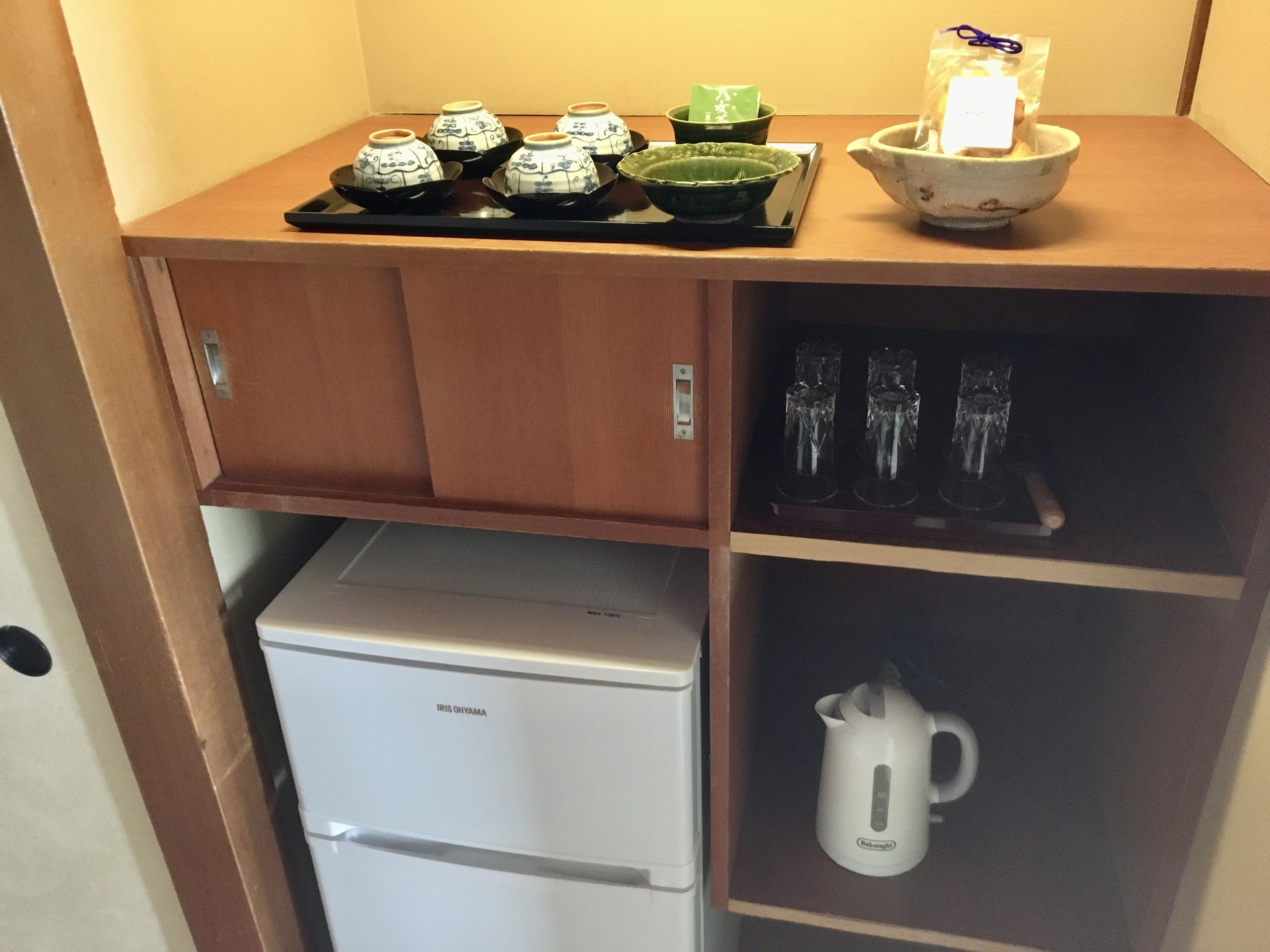 A compact kitchen with a refrigerator, a shelf with teapots, cups, and a kettle.
