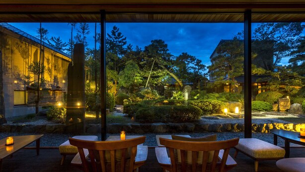 An outdoor seating area with wooden chairs and tables, overlooking a garden with trees and a building in the background. - Hotel Yagi (Awara)