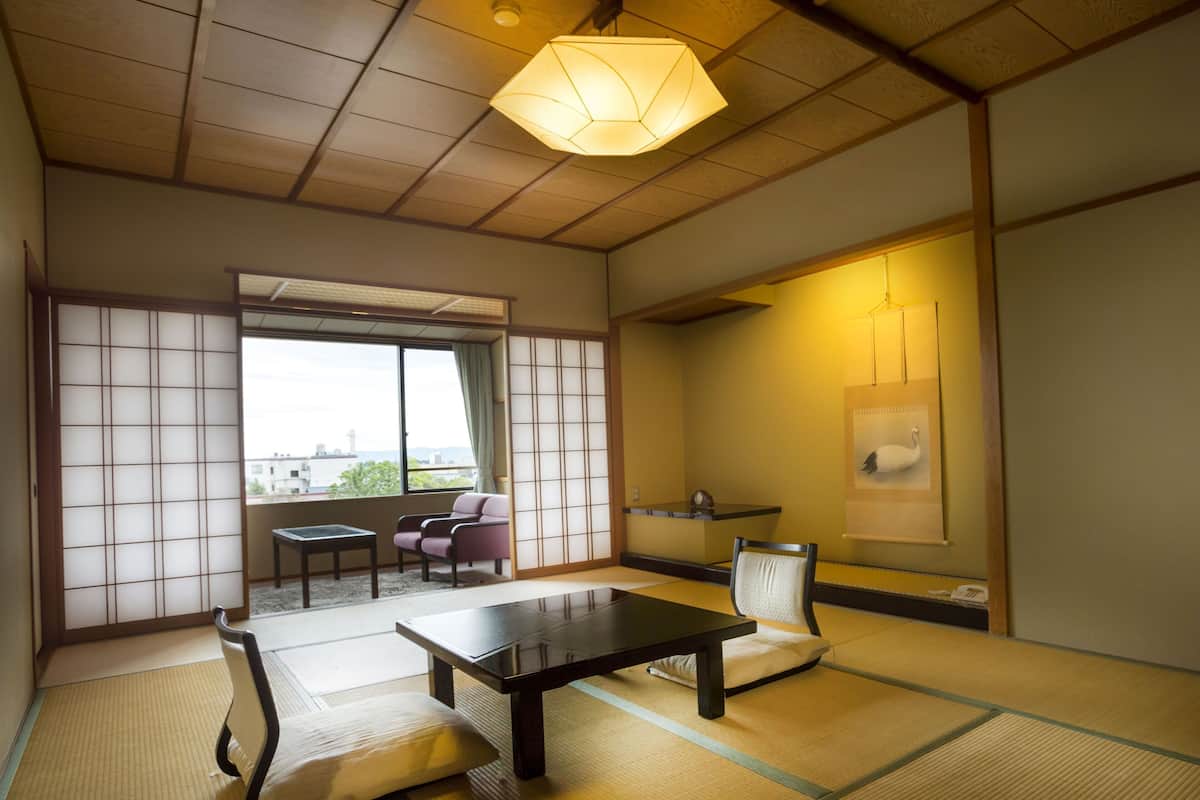 Um quarto japonês tradicional com piso de tatame, uma mesa baixa, uma cadeira, um sofá, uma porta deslizante e vista pela janela.