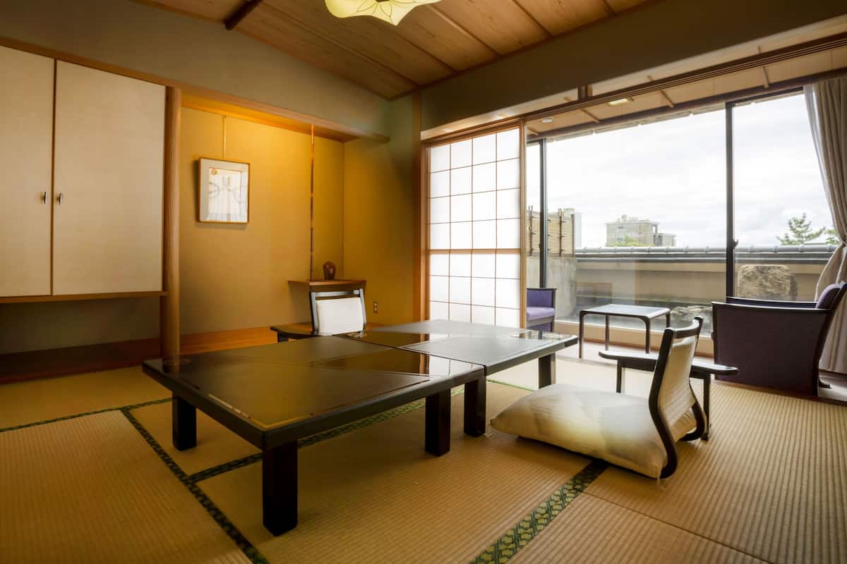 Um quarto japonês tradicional com tatames, uma mesa baixa, uma cadeira estofada e uma grande janela com vista para a cidade.