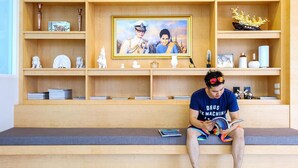 A person sitting on a bench reading a book in a room with wooden shelves displaying various decorative items and a framed picture. - Donchan Residence (Chiang Mai)