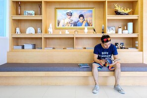 A person sitting on a bench reading a book in a room with wooden shelves displaying various decorative items and a framed picture. - Donchan Residence (Chiang Mai)