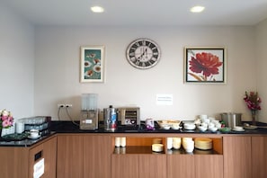 A hotel reception area with a clock, framed floral artwork, a coffee machine, and a buffet counter with stacked plates. - Donchan Residence (Chiang Mai)