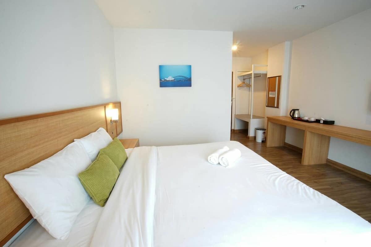 A hotel room with a bed, a bedside table with a kettle, a bench, and a painting on the wall.