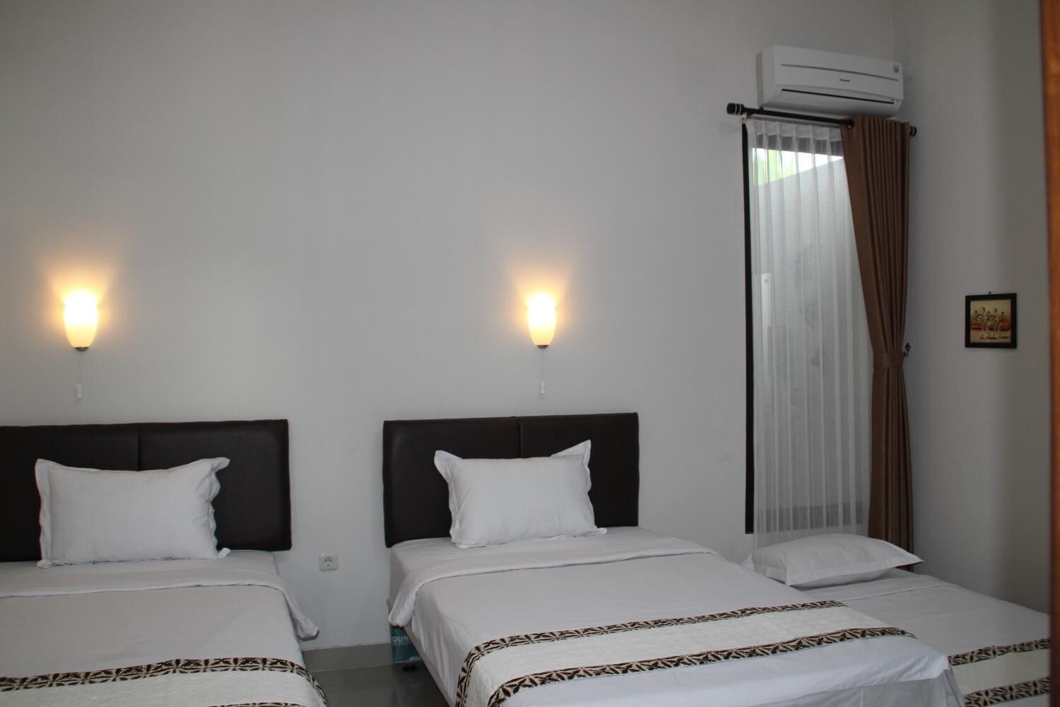 A hotel room with two beds, each with a dark headboard and white bedding. There are wall-mounted lights above each bed, and an air conditioning unit above the window.