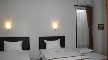 A hotel room with two beds, each with a dark headboard and white bedding. There are wall-mounted lights above each bed, and an air conditioning unit above the window.