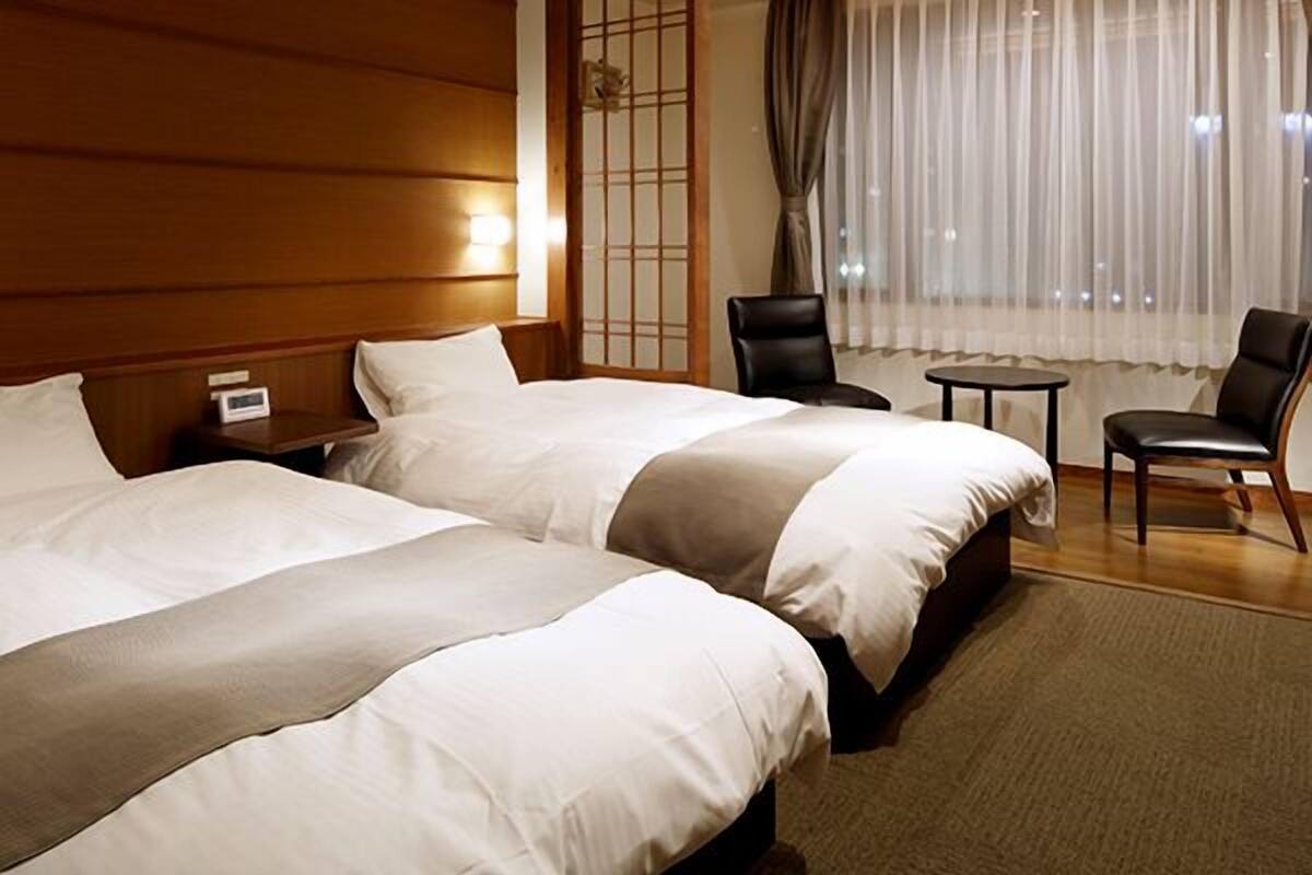 Une chambre d’hôtel avec deux lits, une petite table, deux chaises, une table de chevet et une fenêtre avec des rideaux.