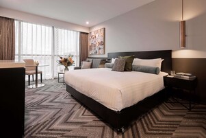 Habitación de hotel moderna con una cama grande, un escritorio, una silla y vistas al exterior a través de amplias ventanas.