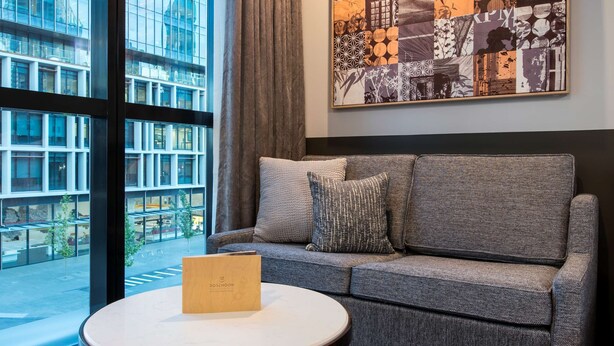 A modern hotel room with a large window, a gray sofa, a round coffee table, and a patterned rug.