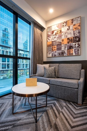 Una habitación de hotel moderna con un ventanal grande, un sofá gris, una mesita de centro redonda y una alfombra estampada.