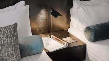 A neatly made bed with white linens, a dark wooden nightstand with a lamp, a magazine, and a glass of water.