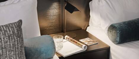A neatly made bed with white linens, a dark wooden nightstand with a lamp, a magazine, and a glass of water.