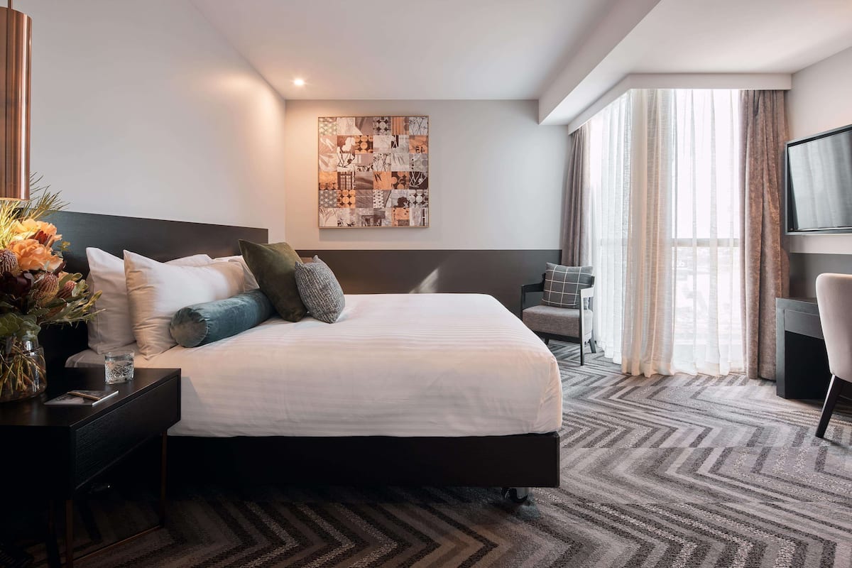 A modern hotel room with a large bed, a desk with a lamp, a flat-screen TV, and a large window with curtains.