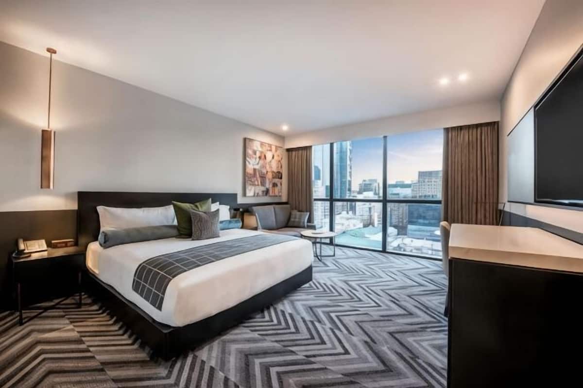 A modern hotel room with a large bed, a desk, a TV, and a city view.