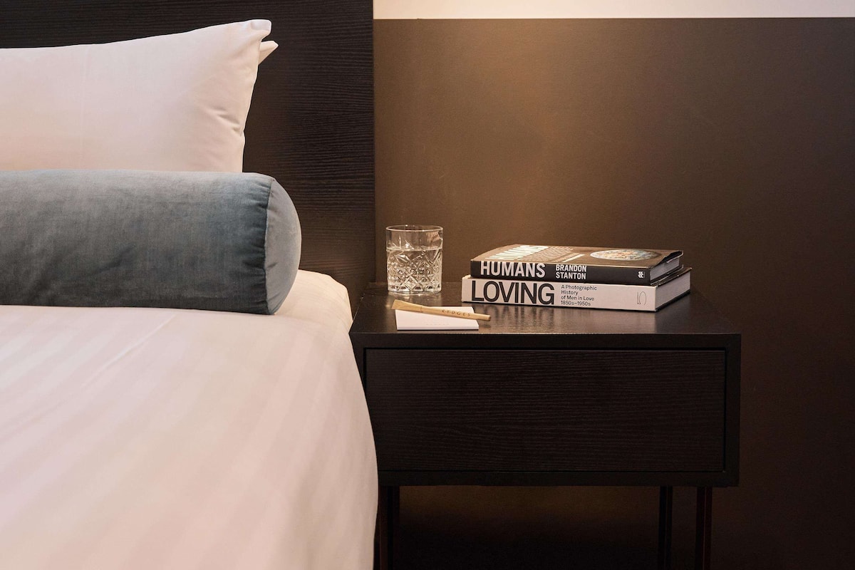 A neatly made bed with pillows, a bedside table with books and a glass, and a wall-mounted light fixture.