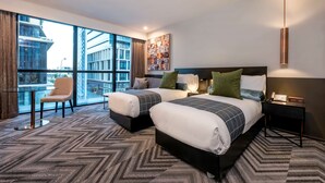 A hotel room with two beds, a chair, a table, and a large window offering a city view.