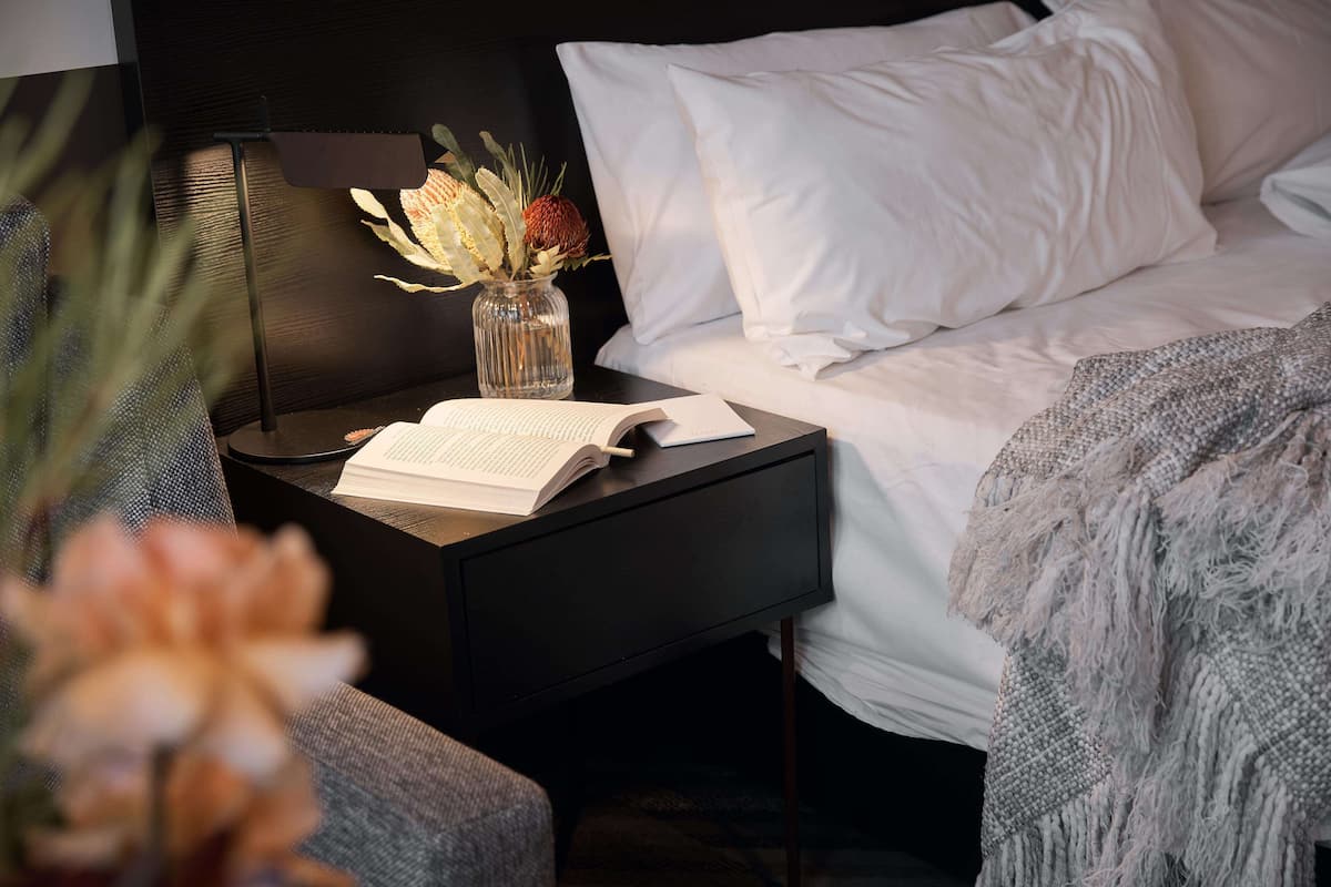 A neatly made bed with white linens, a bedside table with a book and a vase of flowers, and a lamp.