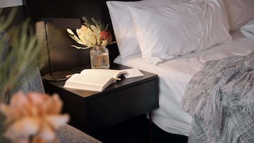 A neatly made bed with white linens, a bedside table with a book and a vase of flowers, and a lamp.