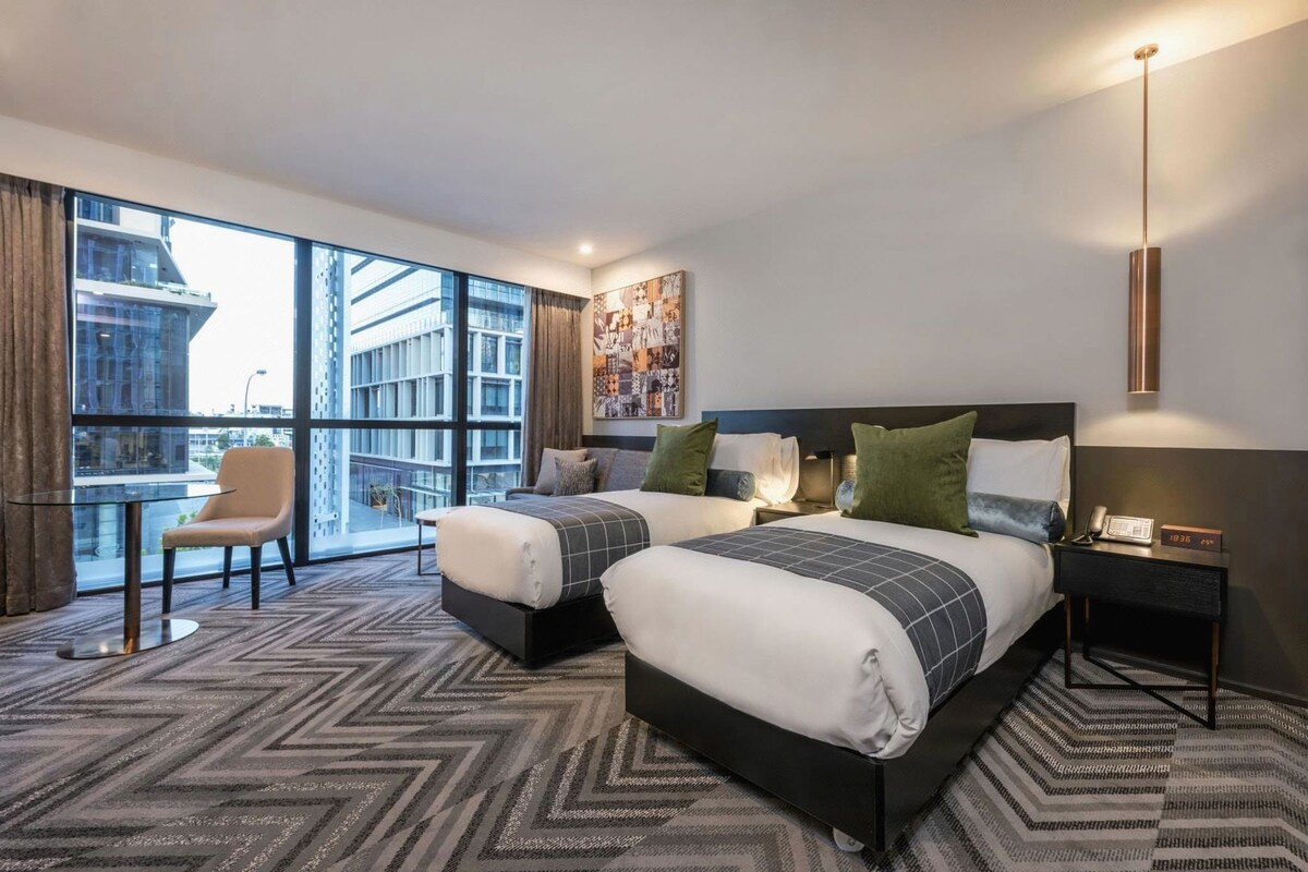 Une chambre d’hôtel avec deux lits, un coin salon avec un fauteuil, une grande fenêtre et une vue sur la ville.