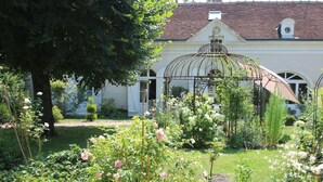 Ein Garten mit verschiedenen Pflanzen und Blumen, einer Pergola und einem Gebäude mit einem Ziegeldach im Hintergrund.