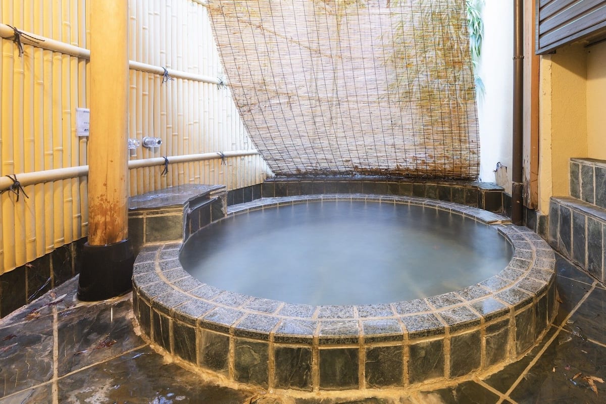 A hot spring bath area with a tiled floor, bamboo wall, and a wire mesh roof.