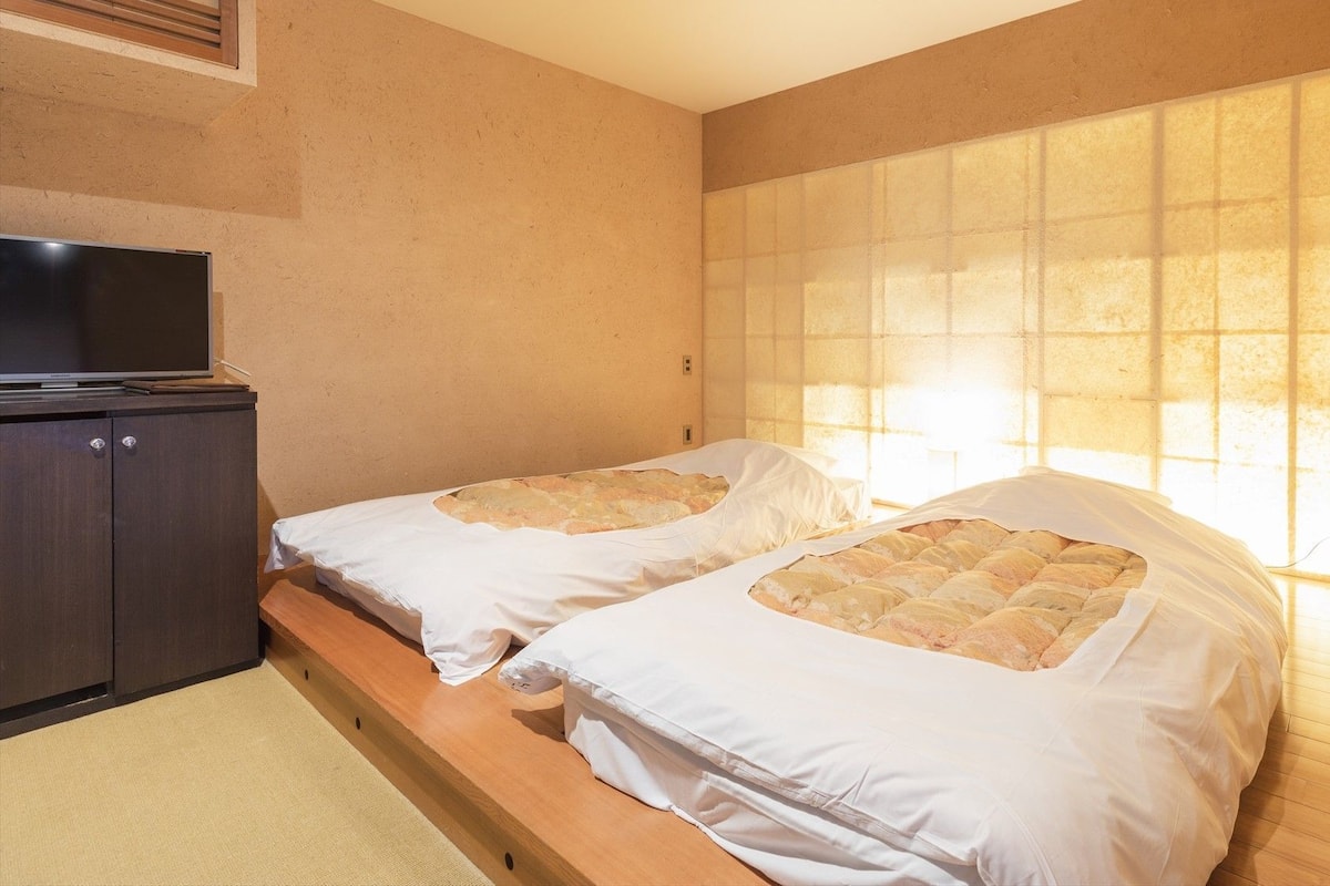 A bedroom with two beds, a TV, and a window with blinds.
